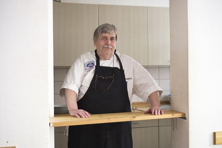 Ein Mann in Schürze, ein Koch, steht an einem Küchentresen oder einer Durchreiche 