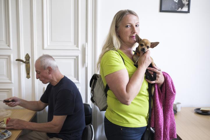 Eine stehende Frau hält einen Hund auf dem Arm und im Hintergrund sitzt ein Mann an einem Tisch