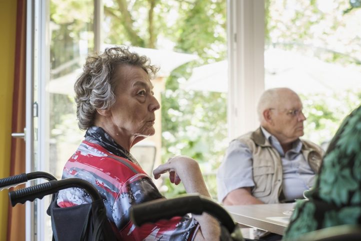 Im Raum eines Pflegeheims sitzt alte Frau im Rollstuhl und guckt zur rechten Seite, im Hintergrund sitze ien alter Mann